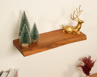 May include: A floating wooden shelf with a warm brown finish, displaying three small green Christmas trees and a gold-colored deer figurine. The shelf is mounted on a white wall, creating a minimalist display.
