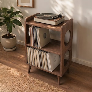 May include: A wooden record player stand with a turntable on top and shelves filled with vinyl records. The stand is a warm brown colour with rounded cutouts on the sides. A potted plant is in the background.