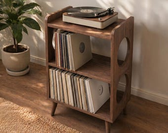 May include: A wooden record player stand with a turntable on top and shelves filled with vinyl records. The stand is a warm brown color with rounded cutouts on the sides. A potted plant is in the background.