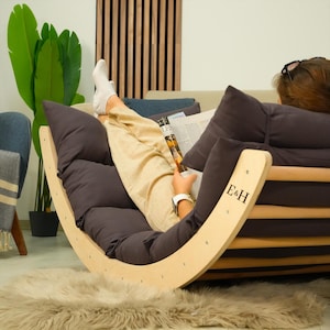 May include: A wooden rocking chair with a dark gray cushion and a person reading a magazine. The chair has a curved design and the letters "E&H" are visible on the side. The person is wearing beige pants and white socks.