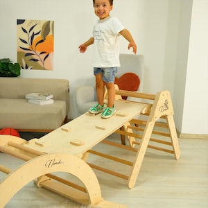 May include: A wooden climbing structure with a ramp and ladder. The ramp has small wooden blocks attached. A child is standing on the ramp. The structure is made of light-colored wood and has the name "Noah" written on it.