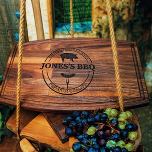 May include: A dark brown wooden chopping board, suspended by rope. The board has the text "JONES'S BBQ" with a pig graphic and the words "SMOKED TO PERFECTION" and "PREMIUM QUALITY" in a circular design. Grapes are in the foreground, with a dark brown finish.