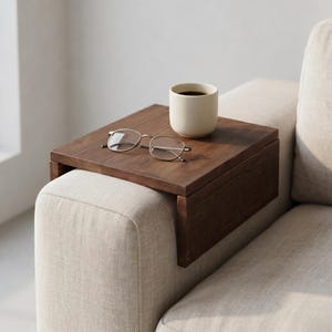 Puede incluir: Una bandeja de brazo de sofá de madera marrón oscuro con una taza de cerámica beige y un par de gafas con montura plateada. La bandeja es rectangular y está diseñada para encajar sobre un brazo de sofá. La taza contiene un líquido oscuro.