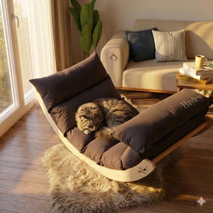 A gray cat is curled up and sleeping on a brown recliner in a cozy living room with a beige sofa, a potted plant, and a window.