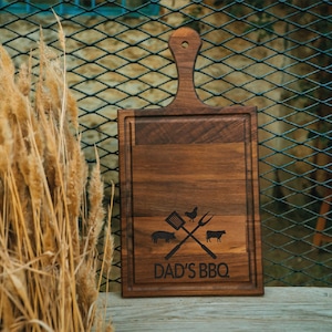May include: A rectangular wooden chopping board with a handle, featuring a carved border and the text "DAD'S BBQ." The design includes a pig, chicken, cow, spatula, and fork.