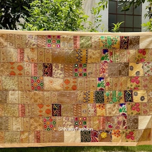 May include: A large, rectangular patchwork textile with a beige border. The patchwork is composed of numerous fabric squares in various shades of gold, brown, and cream, with accents of red, green, and blue. The textile has a decorative, handcrafted appearance.