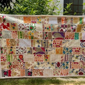 Puede incluir: Un edredón rectangular de patchwork con un borde color crema. El edredón está compuesto por numerosos cuadrados de tela en varios colores y patrones, incluyendo diseños florales y geométricos. El edredón se exhibe al aire libre.