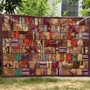 Può includere: Un tessuto patchwork con un bordo marrone, caratterizzato da una varietà di quadrati a motivi nelle tonalità del rosso, oro, verde acqua e marrone. Il tessuto è tenuto contro uno sfondo di fogliame verde ed erba.