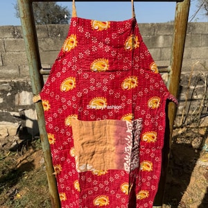 May include: Red apron with a floral and paisley pattern. The apron has a large front pocket with a patchwork design. The apron is hanging on a metal ladder. The text "ShivaayFashion" is visible on the apron.