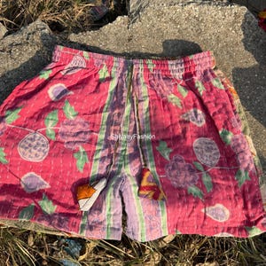 May include: Pink and purple patterned shorts with a drawstring waist. The shorts feature floral and geometric designs, with green and purple vertical stripes. The shorts have decorative charms and the text "ShivaayFashion".