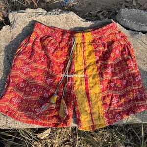 May include: Red and yellow patterned shorts with a drawstring waist. The shorts feature a patchwork design with intricate stitching and a decorative tassel. The fabric has a textured appearance, and the shorts appear to be handmade.