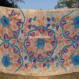 May include: Beige textile with intricate embroidery in shades of blue, orange, and teal. The design features floral and geometric patterns, including stylized flowers and pomegranate shapes. The text "ShivaayFashion" is visible.