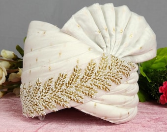 Pode incluir: Um turbante branco com bordado de contas douradas e um design plissado. O turbante é decorado com pequenos pontos dourados e é exibido em uma superfície rosa com flores brancas e vermelhas.