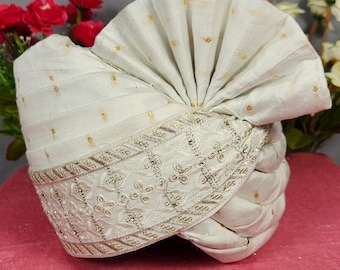 Pode incluir: Um turbante creme com detalhes de pontos dourados e acabamento bordado. O turbante é dobrado e moldado, com um topo decorativo em forma de leque. Ele repousa sobre uma superfície rosa, com flores vermelhas e brancas ao fundo.