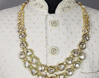 Könnte beinhalten: Ein cremefarbener Sherwani mit texturiertem geometrischem Muster, Stehkragen und runden Knöpfen, verziert mit einer zweireihigen goldfarbenen Halskette aus klaren, unregelmäßig geformten Steinen in einer metallischen Fassung.