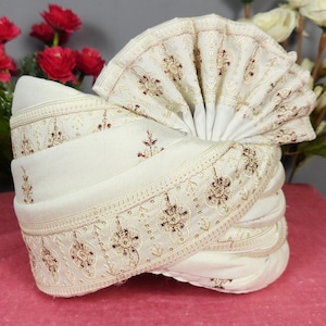 May include: An off-white turban with detailed gold embroidery. The turban is folded and layered, with a fan-shaped element at the top. It rests on a pink surface, with red and white flowers behind. The embroidery includes floral designs.