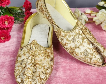 Può includere: Un paio di scarpe slip-on color crema con intricati ricami metallici dorati in un motivo floreale. Le scarpe presentano una punta affusolata, un'apertura scollata e una fodera interna chiara, che mette in risalto la lavorazione dettagliata.