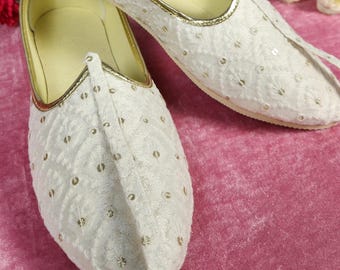 Pode incluir: Um par de sapatos brancos sem atacadores com um intrincado padrão floral bordado e pequenas lantejoulas metálicas. Os sapatos têm uma biqueira pontiaguda, um forro interior amarelo claro e um rebordo dourado na parte superior.