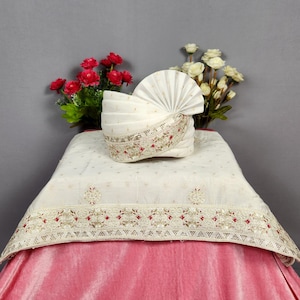 May include: A white turban with detailed embroidery and a pleated fan design, placed on a cream cloth with floral patterns. The cloth is draped over a pink velvet fabric, with red and white flowers in the background.