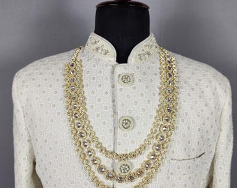 Könnte beinhalten: Cremefarbene, bestickte Jacke mit Stehkragen und goldenen Knöpfen. Eine dreistufige goldene Halskette mit klaren Steinen ist über der Jacke drapiert. Die Jacke wird auf einer Schaufensterpuppe präsentiert.
