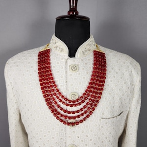 May include: A multi-strand necklace featuring vibrant red beads. The necklace is displayed on a mannequin wearing a white jacket with intricate embroidery and decorative buttons. The beads are a deep red color.