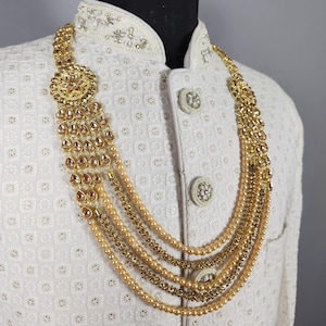 Pu&ograve; includere: Una collana dorata con pi&ugrave; fili e un grande pendente ornato. La collana presenta una combinazione di perline ed elementi decorativi, esposta su una giacca bianca strutturata.