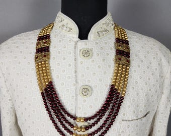 Könnte beinhalten: Eine mehrsträngige Halskette mit goldenen und weinroten Perlen. Die Halskette hat dekorative goldene und weinrote Akzente. Die Halskette wird auf einer Schaufensterpuppe präsentiert, die eine weiße Jacke mit aufwendiger Stickerei und Knöpfen trägt.