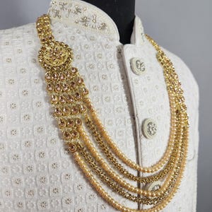 Pu&ograve; includere: Una collana dorata con pi&ugrave; fili e un pendente decorativo con pietre a forma di goccia. La collana &egrave; esposta su una giacca bianca strutturata, mostrando il design e l'artigianato intricati.