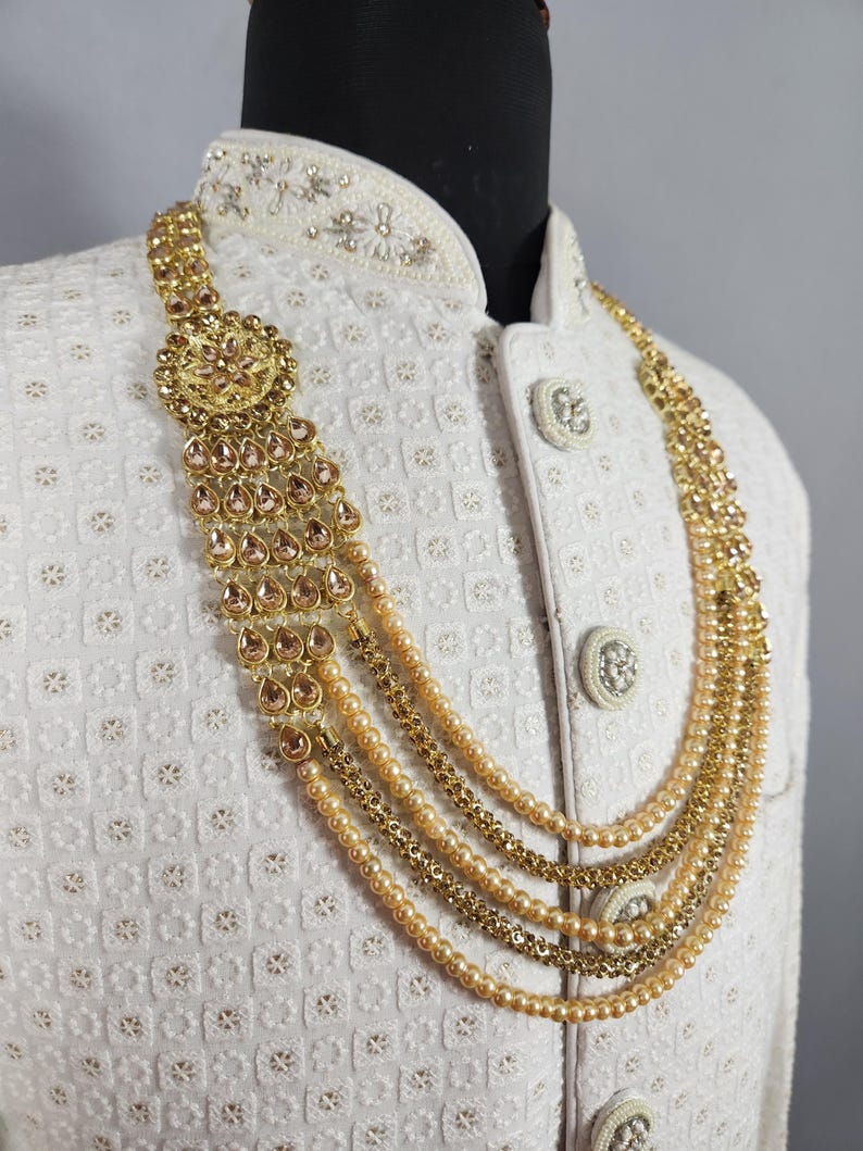 Pu&ograve; includere: Una collana dorata con pi&ugrave; fili e un pendente decorativo con pietre a forma di goccia. La collana &egrave; esposta su una giacca bianca strutturata, mostrando il design e l'artigianato intricati.