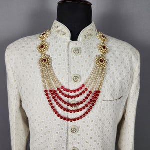 Pu&ograve; includere: Giacca color crema con ricami intricati e collana a strati. La collana presenta perline rosse, perle e dettagli floreali dorati. La giacca ha un motivo strutturato e bottoni decorativi.