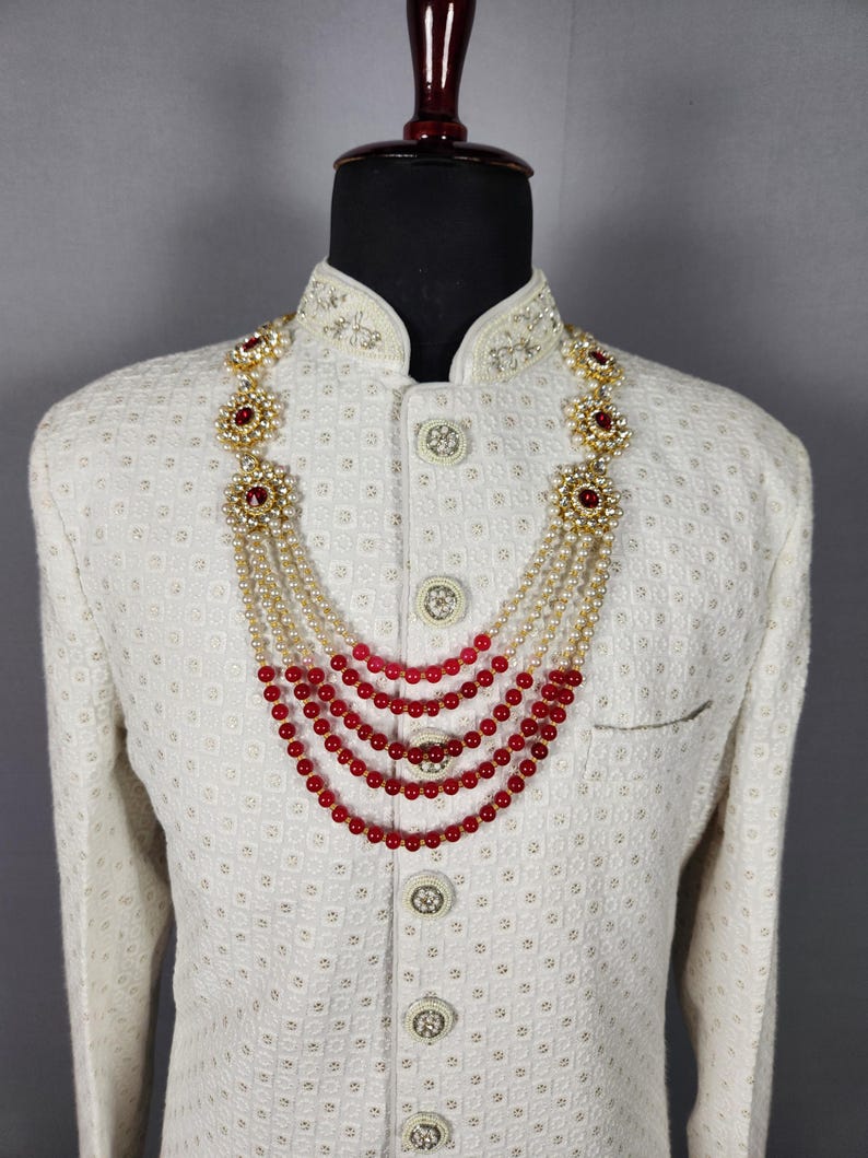 Pu&ograve; includere: Giacca color crema con ricami intricati e collana a strati. La collana presenta perline rosse, perle e dettagli floreali dorati. La giacca ha un motivo strutturato e bottoni decorativi.