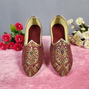 May include: A pair of ornate, pointed-toe shoes with a rich burgundy and gold brocade pattern. The shoes have a gold trim along the opening and a deep burgundy interior. The background features pink fabric and floral arrangements.