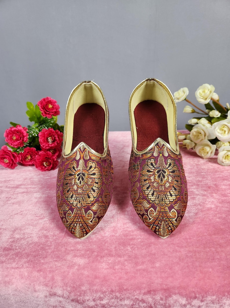 May include: A pair of ornate, pointed-toe shoes with a rich burgundy and gold brocade pattern. The shoes have a gold trim along the opening and a deep burgundy interior. The background features pink fabric and floral arrangements.