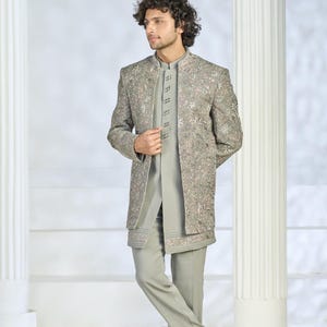 Puede incluir: Un traje bordado gris claro con una chaqueta larga, pantalones y una camisa con cuello mandar&iacute;n. La chaqueta presenta intrincados bordados florales en tonos rosa y plata. El conjunto es formal.
