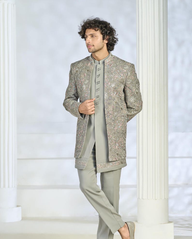 Puede incluir: Un traje bordado gris claro con una chaqueta larga, pantalones y una camisa con cuello mandar&iacute;n. La chaqueta presenta intrincados bordados florales en tonos rosa y plata. El conjunto es formal.