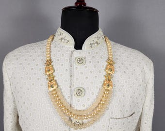 Könnte beinhalten: Eine cremefarbene, dreireihige Perlenkette mit goldfarbenen Akzenten und dekorativen Anhängern. Die Halskette wird auf einer Schaufensterpuppe mit einer weißen Jacke mit aufwendiger Stickerei und Knöpfen präsentiert.