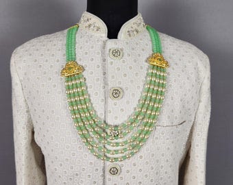 Pode incluir: Casaco creme com bordados intrincados e colar de várias voltas. O colar apresenta contas verdes, pérolas falsas e fechos decorativos dourados. O casaco tem um padrão texturizado e botões ornamentados.