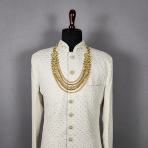 Pu&ograve; includere: Una giacca color crema a maniche lunghe con un sottile motivo geometrico, abbottonata e esposta su un manichino. Una collana d'oro a pi&ugrave; fili &egrave; drappeggiata sulla giacca, aggiungendo un tocco di eleganza. Il manichino ha una testa marrone scuro.