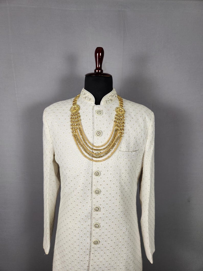 Pu&ograve; includere: Una giacca color crema a maniche lunghe con un sottile motivo geometrico, abbottonata e esposta su un manichino. Una collana d'oro a pi&ugrave; fili &egrave; drappeggiata sulla giacca, aggiungendo un tocco di eleganza. Il manichino ha una testa marrone scuro.