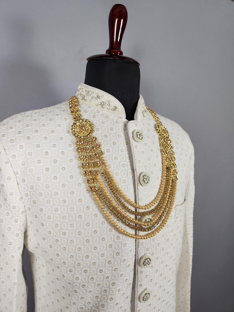 Pu&ograve; includere: Una collana dorata a pi&ugrave; fili &egrave; esposta su un manichino con una giacca bianca. La collana presenta un pendente centrale e strati di perline ed elementi decorativi. La giacca ha un motivo strutturato e bottoni ornati.