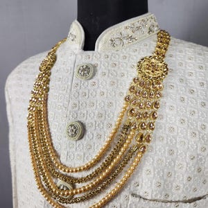 Pu&ograve; includere: Una collana dorata con pi&ugrave; fili di perline ed elementi decorativi. La collana &egrave; a strati e presenta un grande pendente ornato con accenti a forma di goccia. La collana &egrave; esposta su una giacca bianca strutturata con bottoni decorativi.