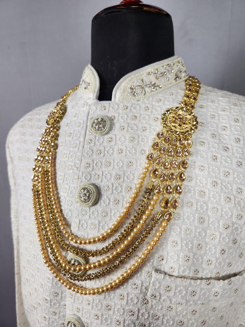 Pu&ograve; includere: Una collana dorata con pi&ugrave; fili di perline ed elementi decorativi. La collana &egrave; a strati e presenta un grande pendente ornato con accenti a forma di goccia. La collana &egrave; esposta su una giacca bianca strutturata con bottoni decorativi.