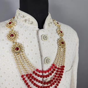 Pu&ograve; includere: Una collana a strati con perline rosse e bianche e dettagli color oro. La collana &egrave; esposta su una giacca bianca con ricami intricati e bottoni decorativi. La collana presenta grandi pietre preziose rosse.