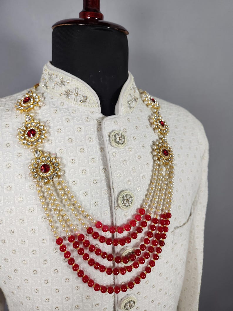 Pu&ograve; includere: Una collana a strati con perline rosse e bianche e dettagli color oro. La collana &egrave; esposta su una giacca bianca con ricami intricati e bottoni decorativi. La collana presenta grandi pietre preziose rosse.