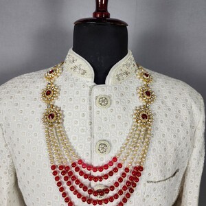Pu&ograve; includere: Una collana a pi&ugrave; fili con fili alternati di perle bianche e perline rosse. La collana presenta accenti floreali color oro con pietre preziose rosse. La collana &egrave; esposta su una giacca bianca con ricami ornati.