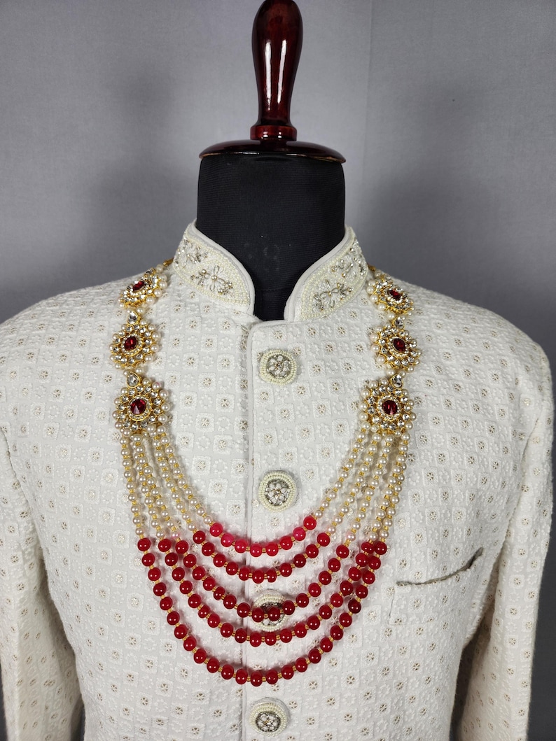 Pu&ograve; includere: Una collana a pi&ugrave; fili con fili alternati di perle bianche e perline rosse. La collana presenta accenti floreali color oro con pietre preziose rosse. La collana &egrave; esposta su una giacca bianca con ricami ornati.