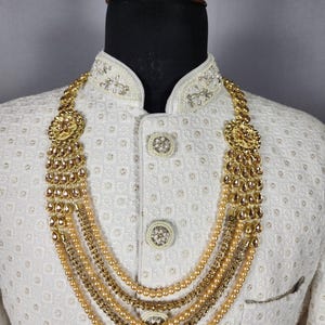 Pu&ograve; includere: Una collana dorata con pi&ugrave; fili di perle ed elementi decorativi. La collana &egrave; esposta su una giacca bianca strutturata con dettagli ornati intorno al colletto e ai bottoni. La collana ha un design a strati.