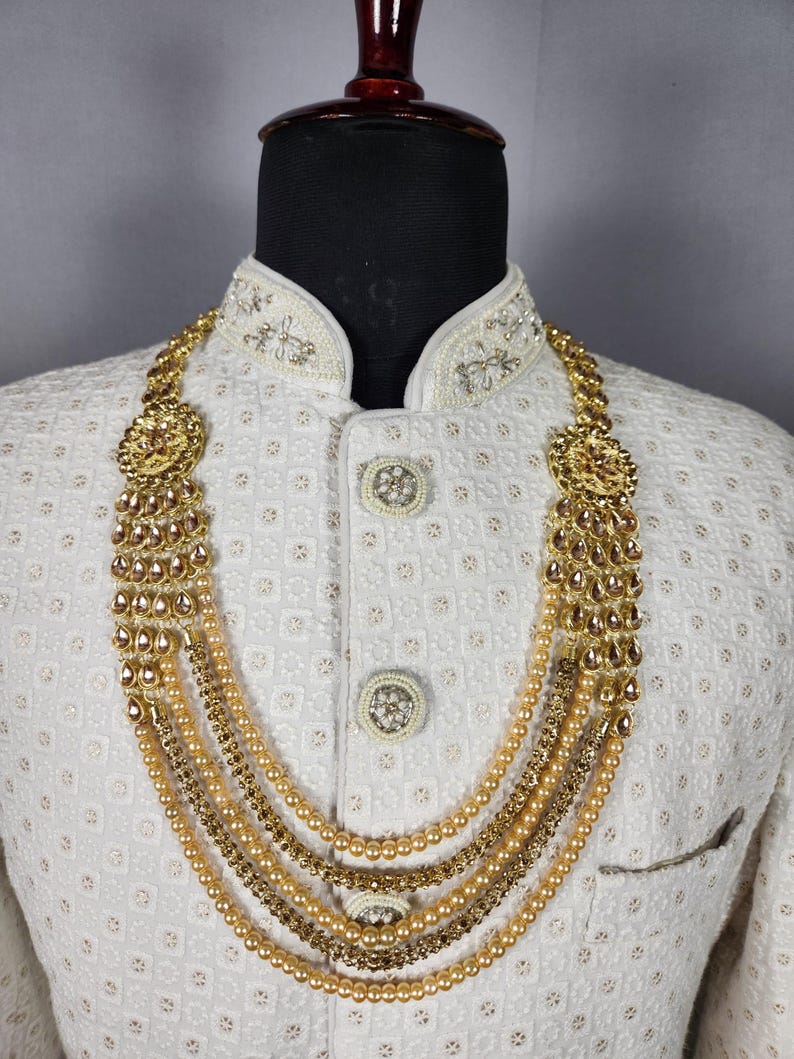 Pu&ograve; includere: Una collana dorata con pi&ugrave; fili di perle ed elementi decorativi. La collana &egrave; esposta su una giacca bianca strutturata con dettagli ornati intorno al colletto e ai bottoni. La collana ha un design a strati.