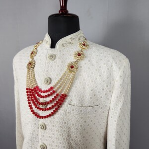 Pu&ograve; includere: Giacca color crema con motivo strutturato e bottoni decorativi. Una collana a pi&ugrave; fili con perline rosse e dettagli floreali dorati adorna la giacca, creando un look formale. La collana presenta pi&ugrave; fili di perline rosse e perle.