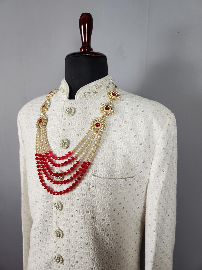 Pu&ograve; includere: Giacca color crema con motivo strutturato e bottoni decorativi. Una collana a pi&ugrave; fili con perline rosse e dettagli floreali dorati adorna la giacca, creando un look formale. La collana presenta pi&ugrave; fili di perline rosse e perle.